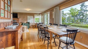 A spacious dining area with a long wooden table, black chairs, and a view of the outdoors through large windows. - Maxwelton Aerie | Bluffside Retreat w/ Sweeping Puget Sound Views + Firepit (Clinton)
