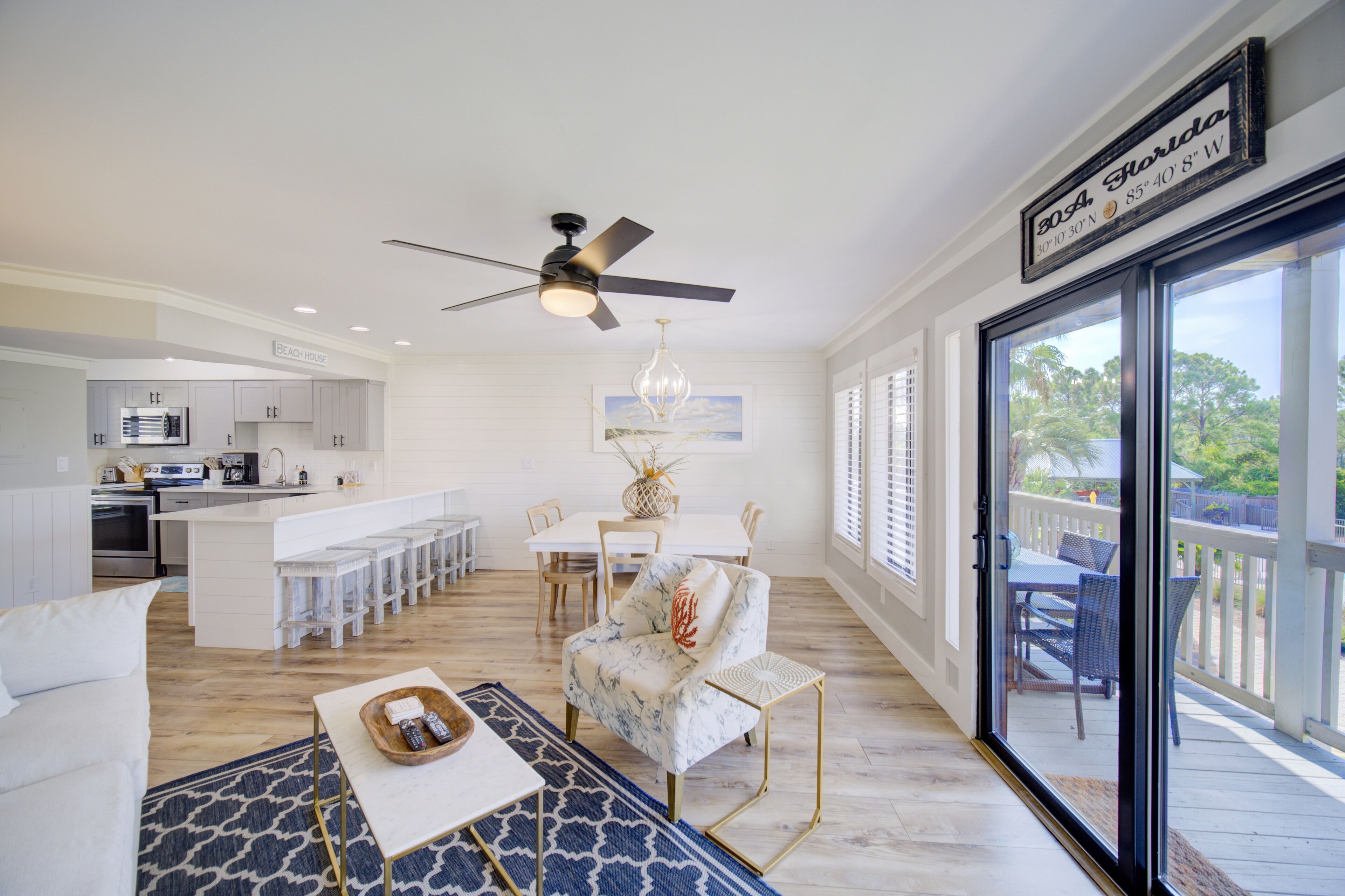 A spacious living area with a white sofa, a coffee table, and a dining table with chairs. There is a kitchen area with white cabinets and a bar. The room has a large window with a view of the outdoors.