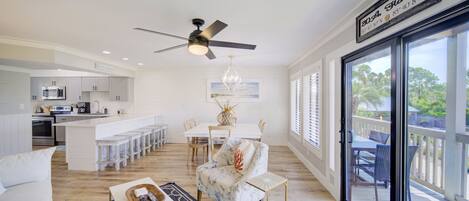 A spacious living area with a white sofa, a coffee table, and a dining table with chairs. There is a kitchen area with white cabinets and a bar. The room has a large window with a view of the outdoors.