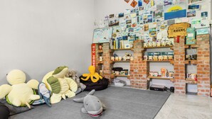 Una habitación con pared de ladrillo, una estantería llena de libros y objetos decorativos, juguetes suaves en el suelo y una pequeña mesa con varios objetos.