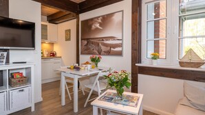 A cozy room with a TV, a dining table with chairs, a small white table with a mat and flowers, and a window with a view of the beach.