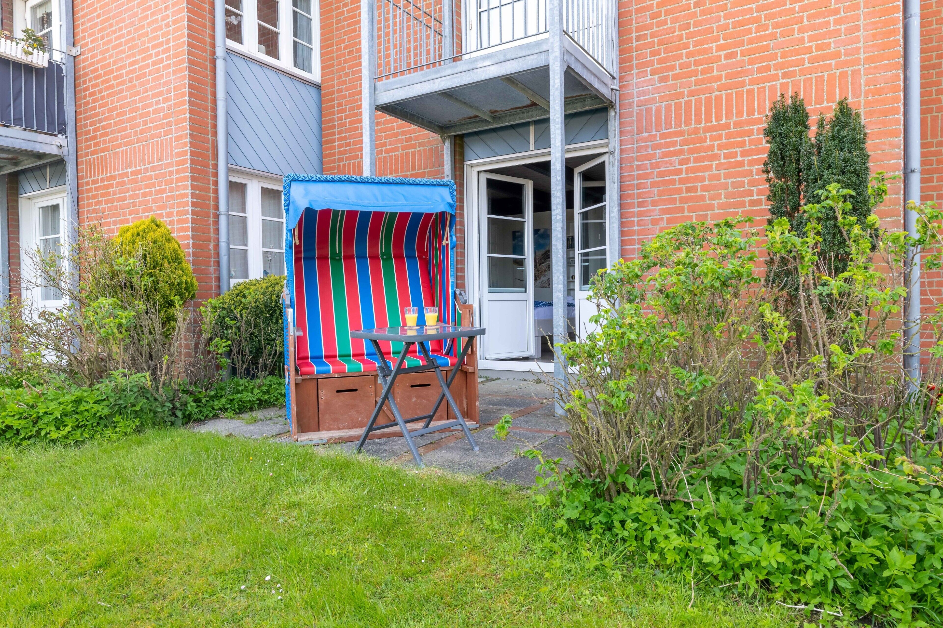 A brick building with a colorful striped awning, a door, and a small garden area with grass and bushes.