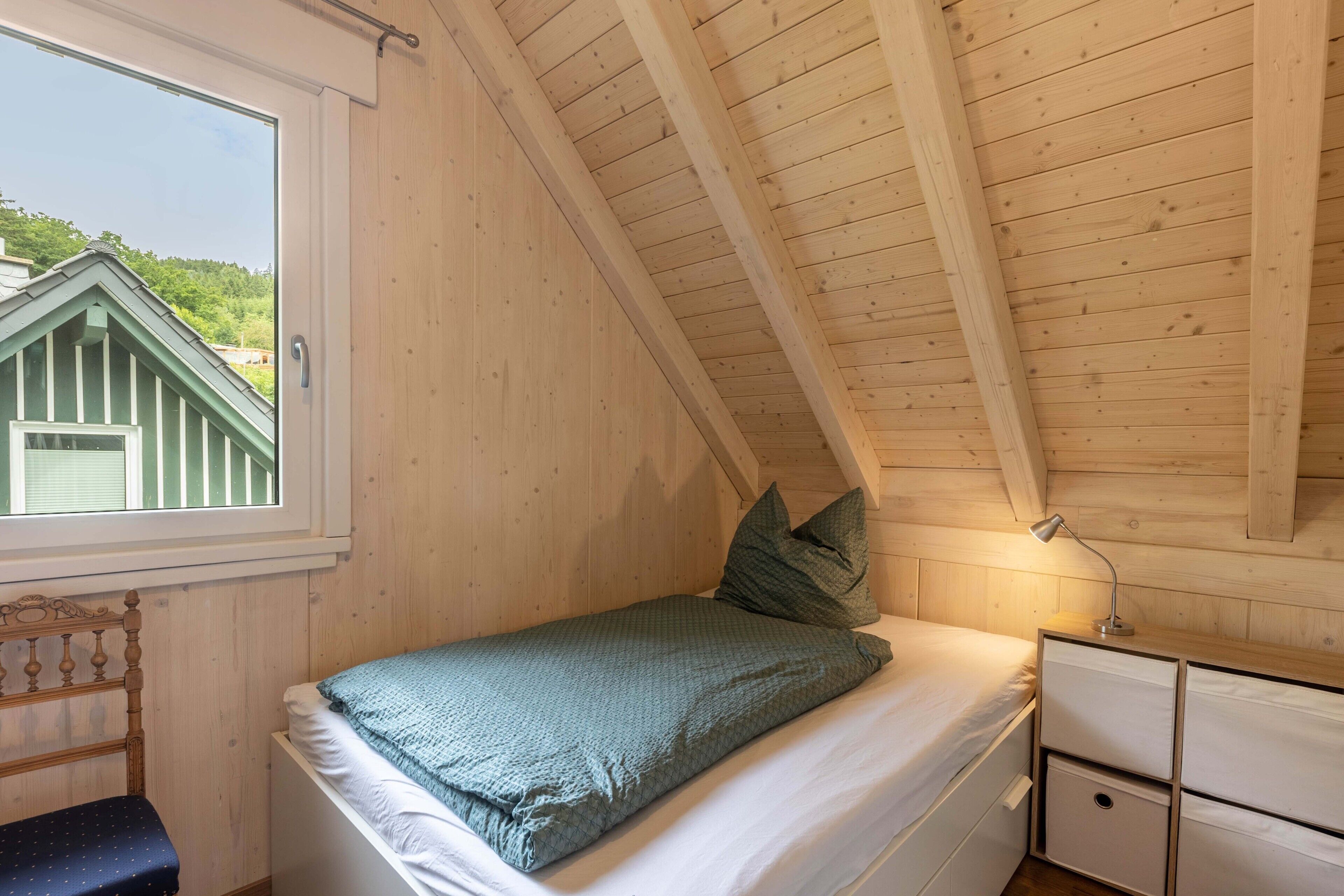A bedroom with a sloped ceiling, a bed with a green blanket, a bedside table with a lamp, and a window with a view of a green building.