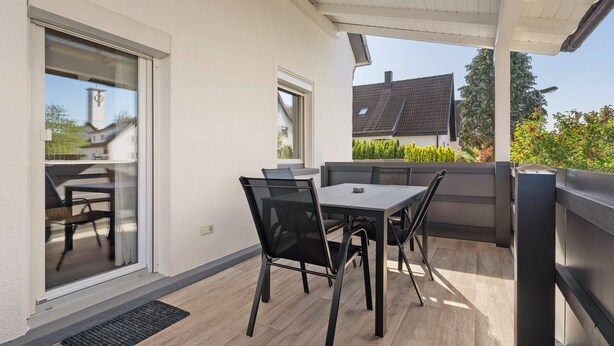 Un patio techado con mesa y sillas, una puerta corrediza de vidrio y vistas a una casa y árboles.