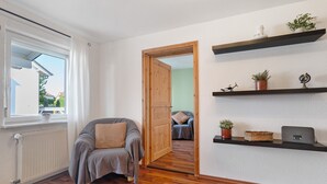 Una habitación con suelo de madera, un sofá gris, un radiador blanco, una puerta de madera y estanterías con plantas y una lámpara.