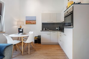 Une cuisine compacte avec des meubles blancs, une table à manger avec des chaises, une lampe et un tableau accroché au mur.