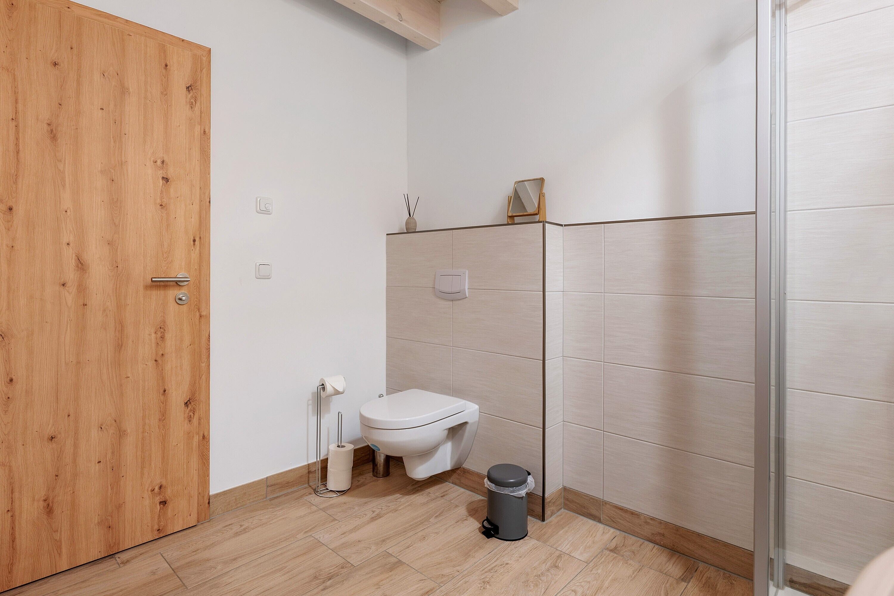 Een badkamer met een houten deur, een toilet, een prullenbak en een douchegedeelte.
