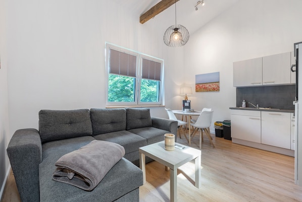 Un salon moderne avec un canapé gris, une petite table blanche et un coin cuisine doté de meubles blancs et d’un plan de travail gris.