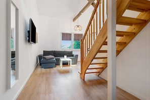 Un salon moderne avec un escalier en bois, un téléviseur à écran plat fixé au mur, un canapé gris, une petite table blanche et une fenêtre avec des stores.