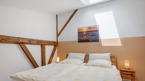 Une chambre à coucher avec un lit, des poutres en bois, une table de chevet, une lampe fixée au mur et un tableau représentant un coucher de soleil.