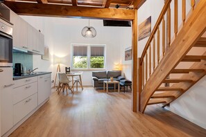 A modern kitchen with white cabinets, a dining area with a table and chairs, a sofa, and a wooden staircase.