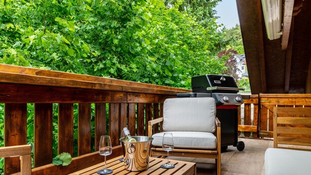 Una terraza de madera con una parrilla, una silla acolchada y una mesa con un balde de vino y copas.