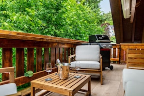 Una terraza de madera con una parrilla, una silla acolchada y una mesa con un balde de vino y copas.
