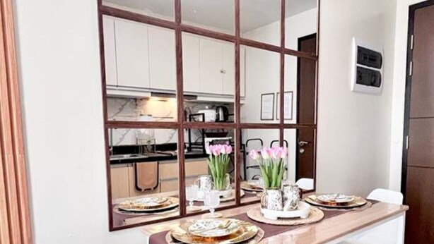 Un comedor con una mesa de madera, sillas blancas, un espejo y una cocina al fondo.
