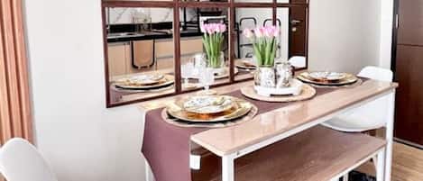 Uma sala de jantar com mesa de madeira, cadeiras brancas, um espelho e uma cozinha ao fundo.