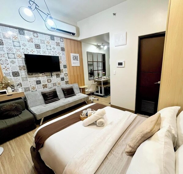 A hotel room with a bed, a sofa, a TV mounted on the wall, and a desk with a chair. - The B's Crib Saint Dominique (Iloilo City)