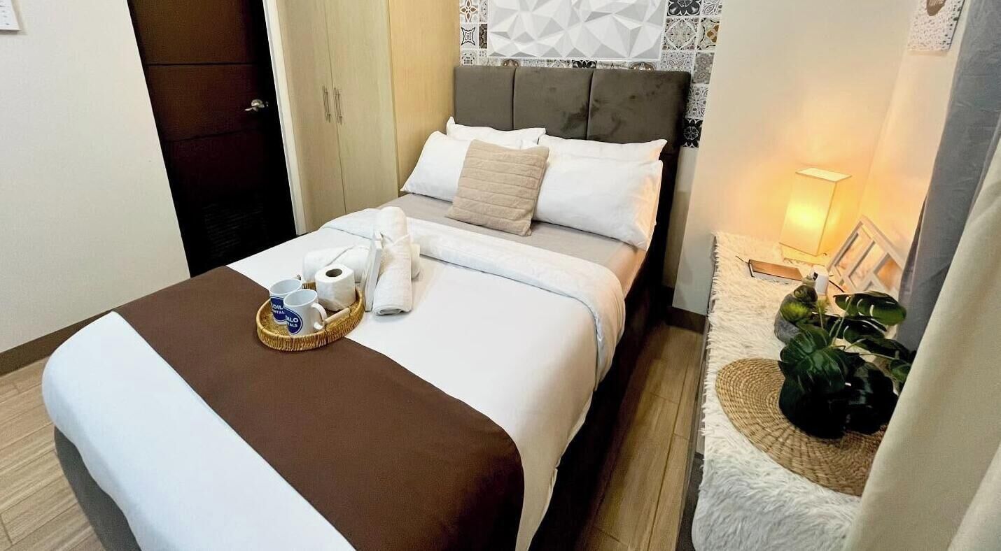 A hotel room with a bed, a bedside table with a lamp, a plant, and a tray with toiletries.