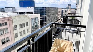 Un balcón con una pequeña mesa y sillas, vista panorámica de la ciudad y una zona ajardinada.