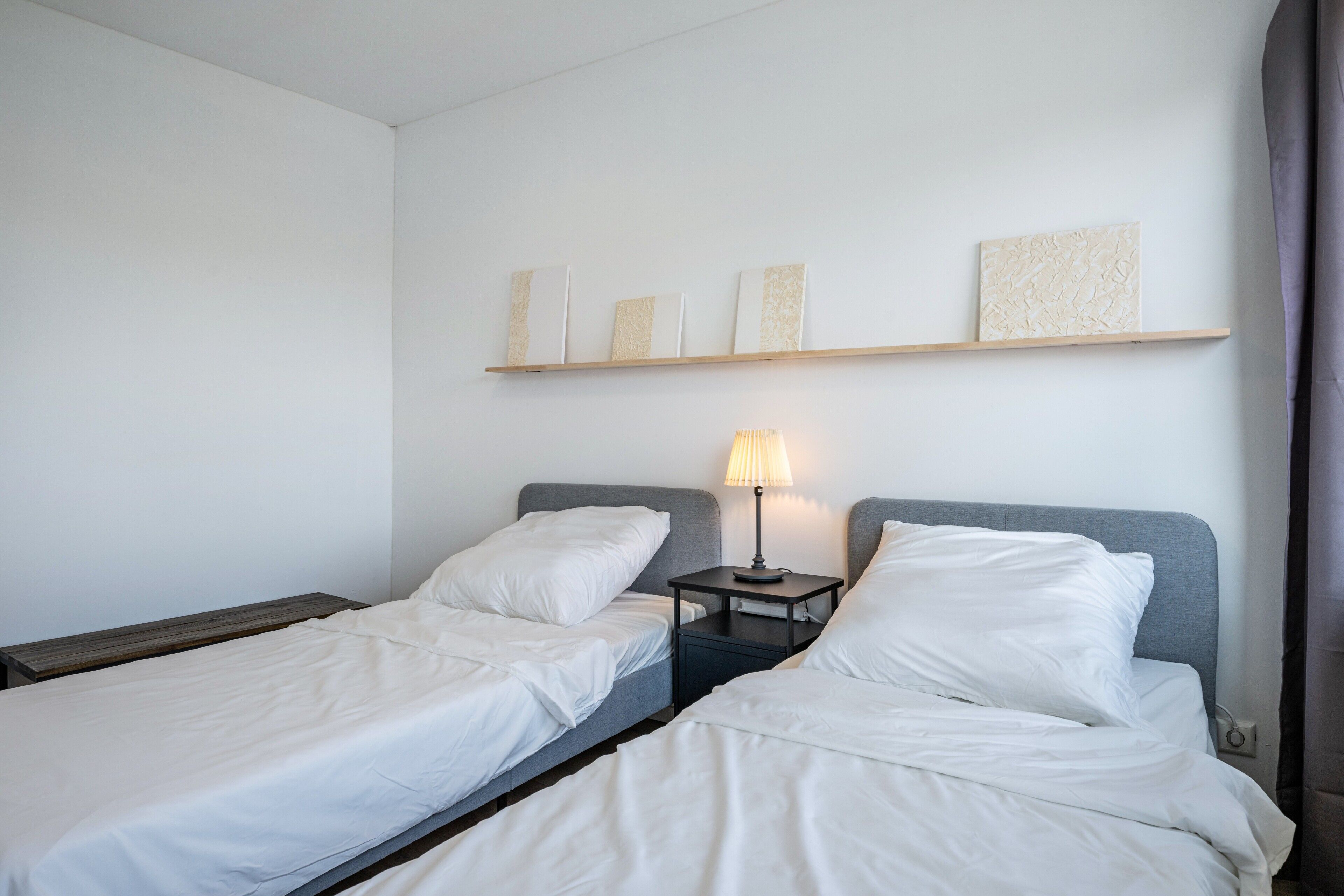 A hotel room with two single beds, a nightstand with a lamp, and a shelf with decorative items.