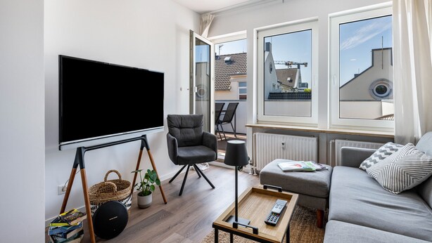 A modern living room with a flat-screen TV, a grey sofa, a black armchair, a wooden coffee table, and a large window with a view of buildings. - Apartment 'Mit Dachterrasse' with Lake View, Balcony and Wi-Fi (Heide)