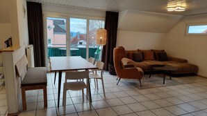 A living room with a sofa, a dining table with chairs, a bench, and a large window. - Apartment '5' with Lake View, Shared Terrace and Wi-Fi (Lindau am Bodensee)