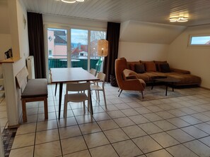 Een woonkamer met een bank, een eettafel met stoelen, een bankje en een groot raam.