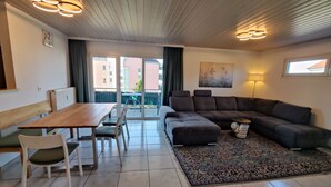 Un salon moderne avec une table à manger en bois, des chaises blanches, un canapé d’angle foncé, un tapis à motifs et un plafond orné de luminaires circulaires.