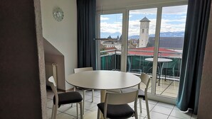 Een kamer met een ronde tafel en vier stoelen, grote ramen met uitzicht op een toren en bergen, en een klok aan de muur.