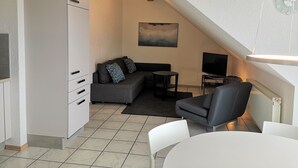 Een moderne loft-achtige woonruimte met kitchenette, een grijze bank, een klein rond tafeltje, een televisie en een witte eettafel.
