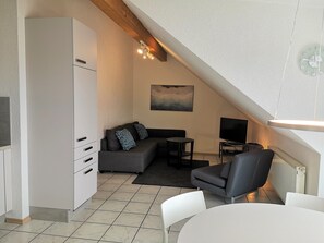 Ein moderner Loft-Wohnraum mit Küchenzeile, einem grauen Sofa, einem kleinen runden Tisch, einem Fernseher und einem weißen Esstisch.