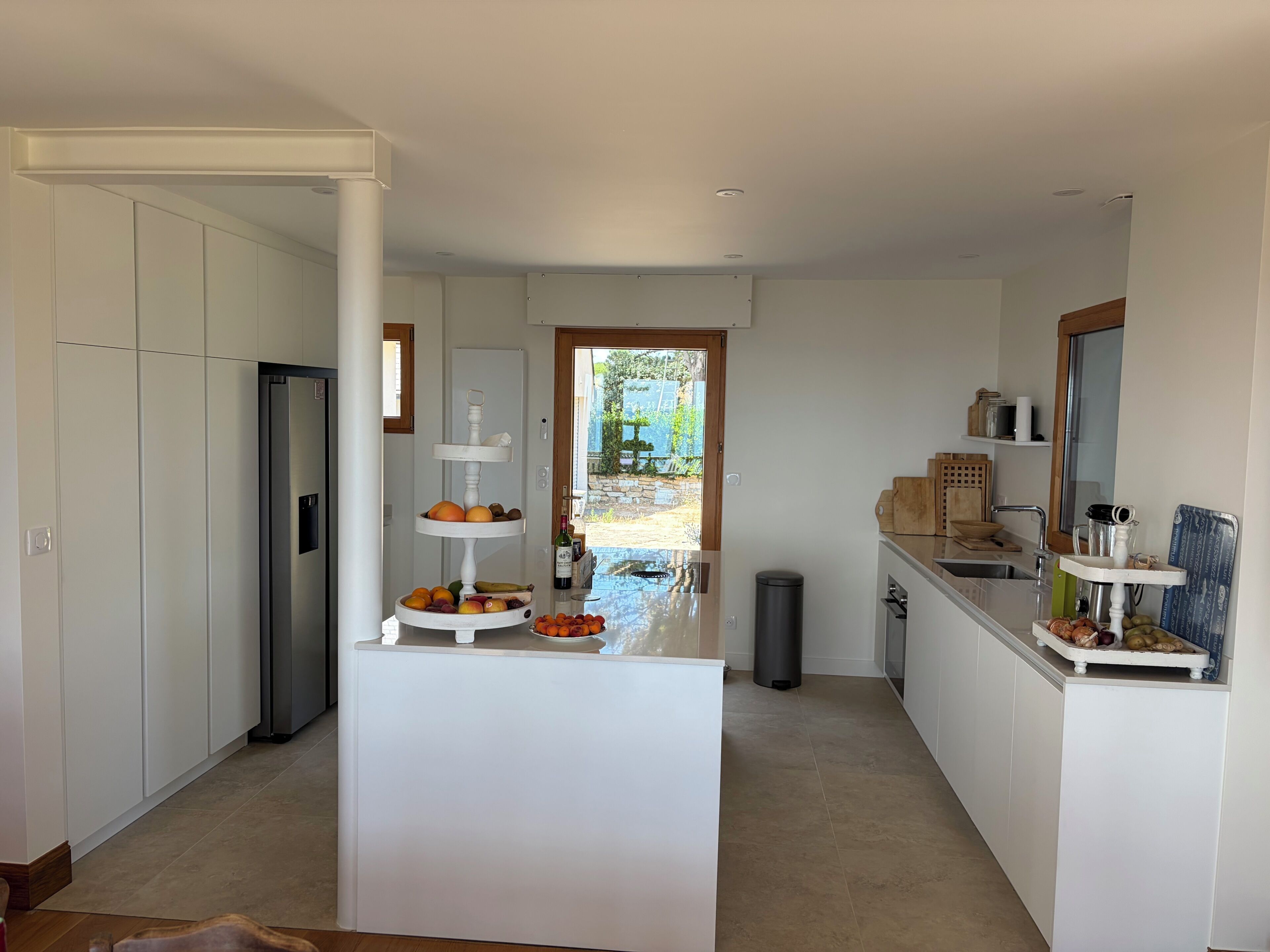 Une cuisine moderne avec des meubles blancs, un îlot central agrémenté de fruits, un évier et une vue sur l’extérieur.
