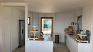 A modern kitchen with white cabinetry, a central island with fruit, a sink, and a view to the outside.