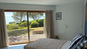 A bedroom with a bed, a view through a sliding door to a garden, a framed picture on the wall, and a door with a handle.