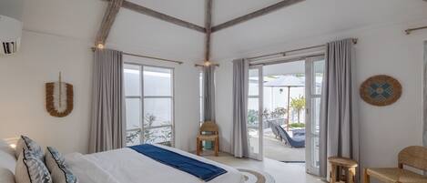 A spacious bedroom with a large bed, wooden beams on the ceiling, and a view of the outdoors through large windows.