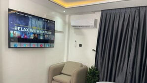 A modern hotel room with a flat-screen TV displaying a nature-themed video, a comfortable armchair, a neatly made bed, and an air conditioning unit. - Kayla City (Kumasi)