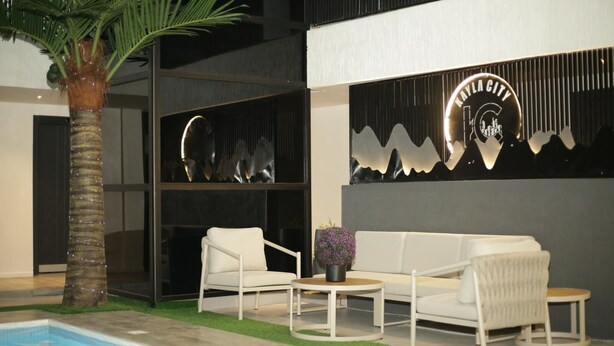 A modern indoor pool area with a white sofa set, a small table, and a palm tree. - Kayla City (Kumasi)