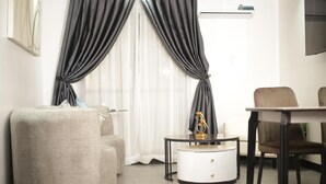A modern living room with a sofa, a round coffee table with a decorative item, a dining table with chairs, and a large window with curtains. - Kayla City (Kumasi)