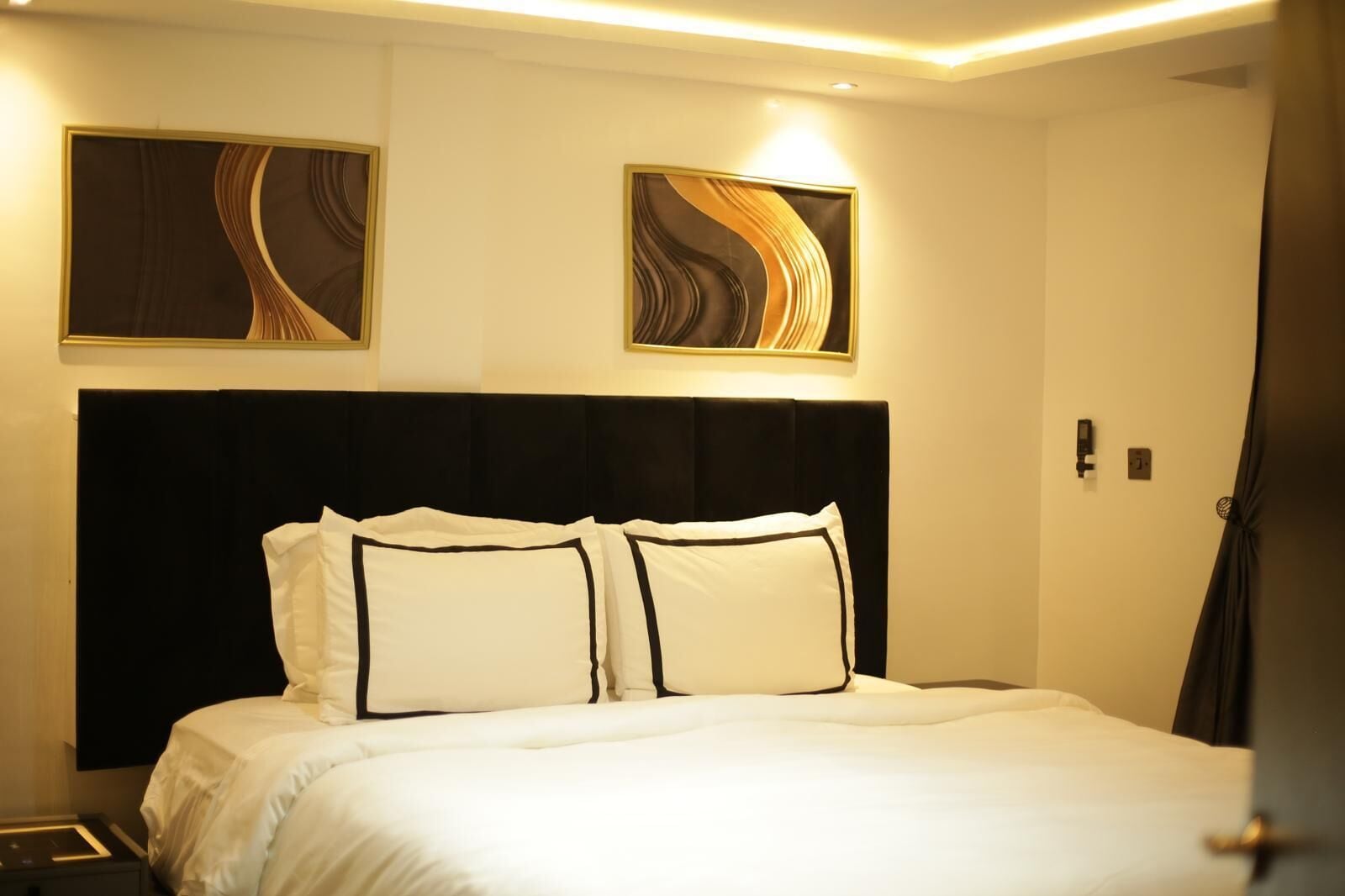 A hotel room with a large bed, two wall-mounted artworks, and a dark headboard.