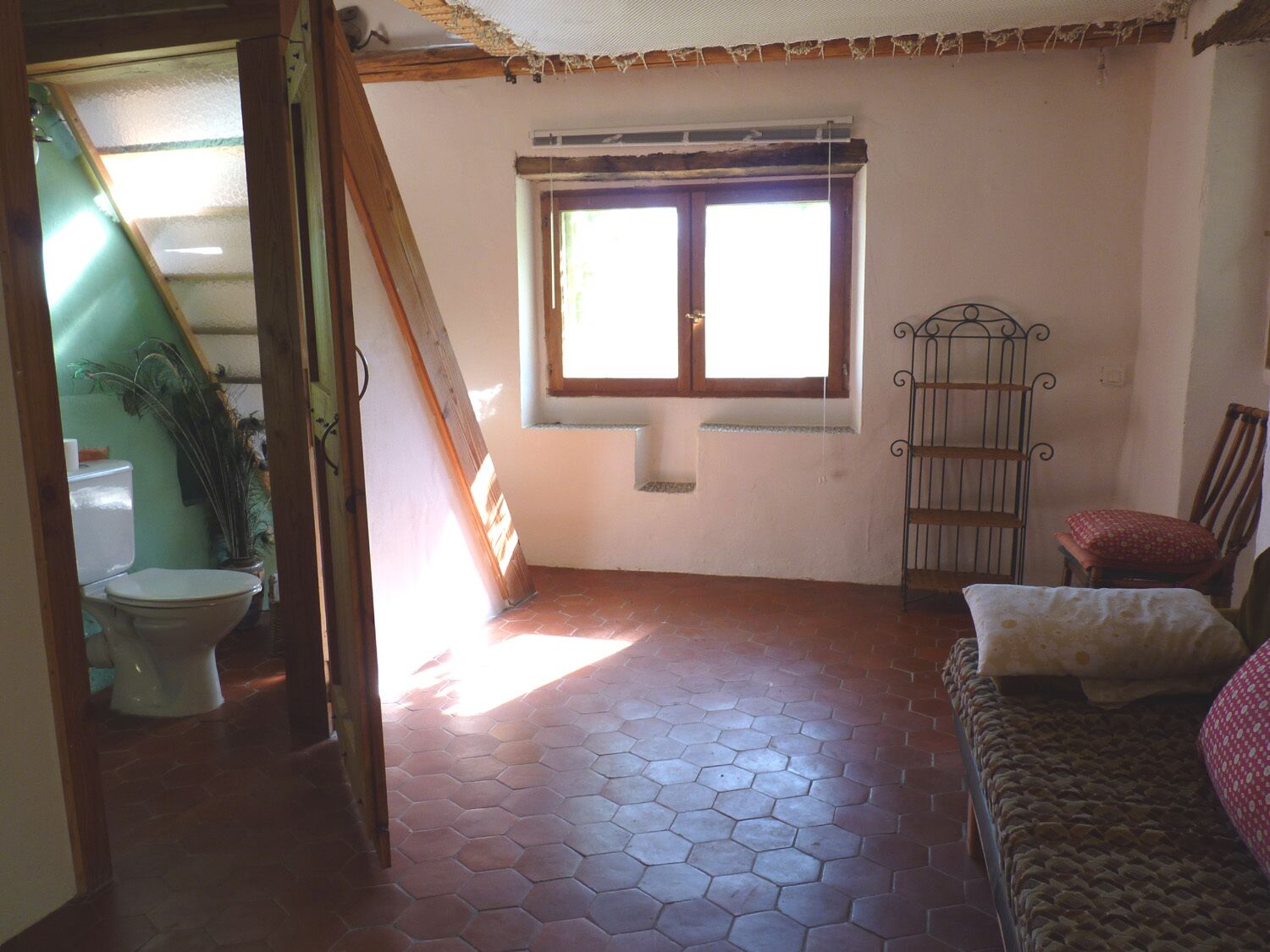 A room with a hexagonal tiled floor, a window with wooden frames, a metal chair, a wooden bed, and a toilet visible through an open door.
