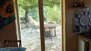 Cuina amb fornel, rentapelats i una paret decorada amb rajoles. A l'exterior, hi ha un patell amb caires i taula, envoltat de verdura.