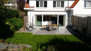 A two-story house with a red-tiled roof, a patio with outdoor furniture, and a sliding glass door leading to an interior space. - Dunette - Beautiful 3-bedroom villa with all comforts in the concession. (De Haan)