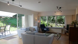 A modern living room with a sofa, a chair, a coffee table, and a TV stand. There is a large window with a view of greenery outside. - Dunette - Beautiful 3-bedroom villa with all comforts in the concession. (De Haan)
