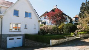 A white two-story house with a garage, surrounded by well-maintained gardens and a clear sky. - Dunette - Beautiful 3-bedroom villa with all comforts in the concession. (De Haan)