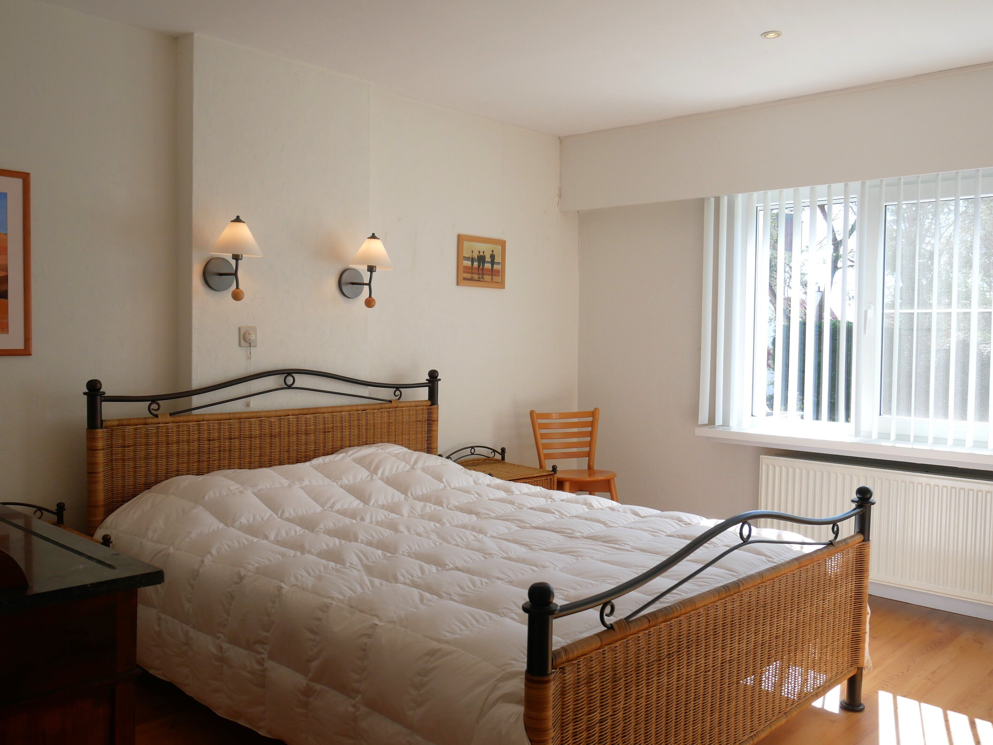 A bedroom with a bed, two wall-mounted lamps, a chair, and a window with blinds.