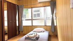 A room with wooden walls, a bed with a white mattress and pillows, a window with curtains, and an air conditioning unit. - coco stay ishigaki (Ishigaki)
