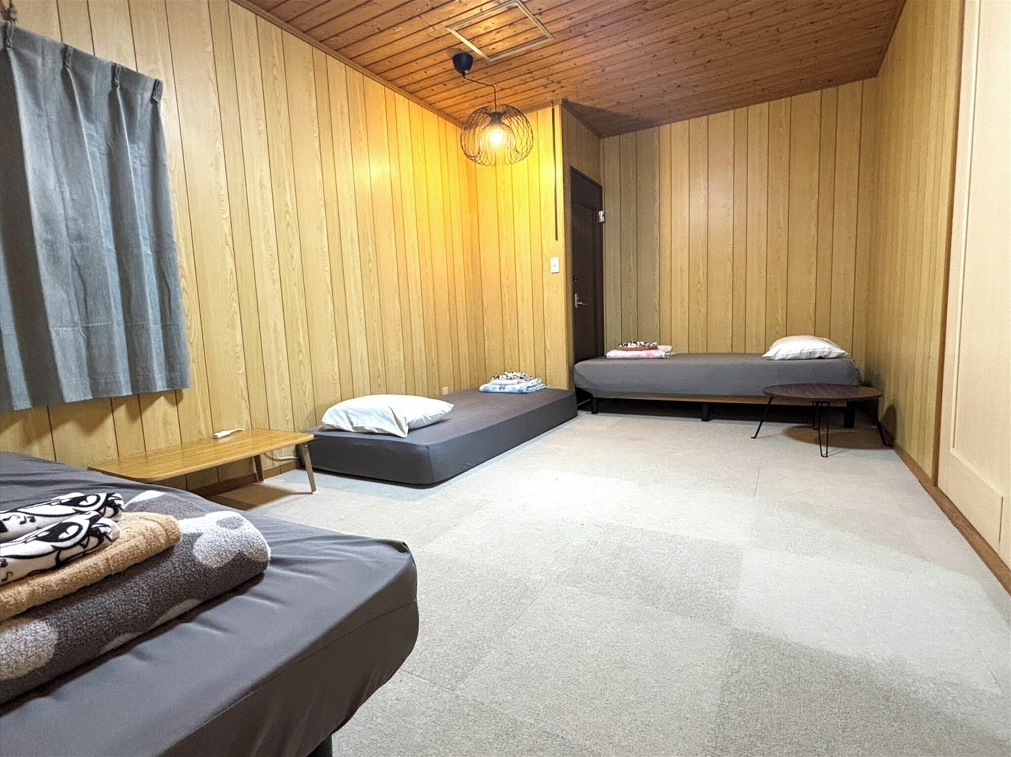 A room with wooden walls, a ceiling light, two beds with pillows, a bench, and a small table.