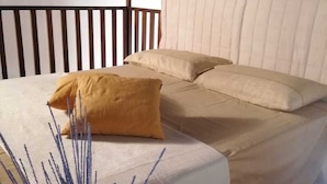 Cama com cabeceira bege e almofadas, corrimão de madeira e uma planta ornamental com flores roxas.