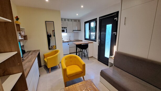 A modern living room with a sofa, two yellow chairs, a wooden coffee table, and a kitchen area with cabinets and a dining table. - Studio Cécilia (Aix-les-Bains)
