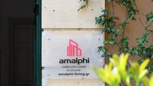Rakennuksen sisäänkäynti, jossa on sininen kyltti, jossa lukee numero 46, sekä kyltti, jossa on teksti »amalphi», logo ja kuvaus: »CORFU CITY LIVING, 2. KERROS».
