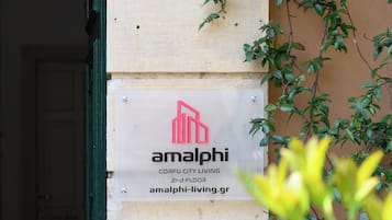 Ingresso di un edificio con un cartello blu che indica il numero 46 e una targa per 'amalphi' con un logo e testo che lo descrive come 'CORFU CITY LIVING 2° PIANO'.